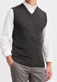 Gilet sans manches gris foncé avec un col en V, une encolure et un ourlet côtelés, porté sur une chemise blanche à manches longues. Tissu en mélange de coton.