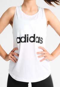 Camiseta de tirantes blanca con un gran logo negro de "adidas", diseño sin mangas, tela ligera, ajuste holgado, usada sobre un sujetador deportivo azul verdoso.