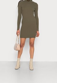 Olivegrön ribbad klänning med långa ärmar och hög krage, kombinerad med en ljusbeige mikro väska och krämfärgade ankelstövlar. Texturen tillför visuell intresse.