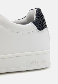 Sneaker bianco in pelle con dettaglio intrecciato blu navy sul tallone, superficie liscia e suola in gomma stampata con marchio in rilievo.