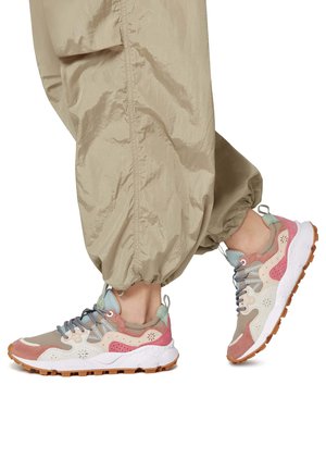Person trägt beige Jogginghosen und mehrfarbige Sneakers mit weißen Sohlen und Gummiprofil, steht auf Zehenspitzen.