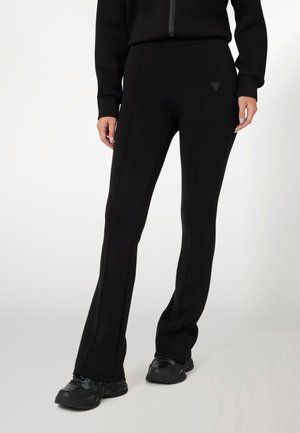 Personne portant un pantalon flare noir ajusté avec des coutures et des baskets noires, debout devant un fond clair uni.