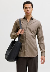 Καφέ πουκάμισο με κουμπιά και μακριά μανίκια, συνδυασμένο με σκούρο γκρι παντελόνι. Κρατά μια μαύρη δερμάτινη τσάντα ώμου με δομημένο σχέδιο και ρυθμιζόμενο ιμάντα.