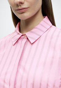 Ροζ πουκάμισο με γιακά και κάθετες λευκές ρίγες, με κλείσιμο κουμπιών στον λαιμό. Ομαλή υφή, εφαρμοστή γραμμή και κλασικός σχεδιασμός.