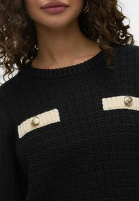 Zwarte gebreide trui met crèmekleurige strepen en gouden decoratieve knopen op de bovenste voorkant. Gelaagd stof en geribbelde halslijn.