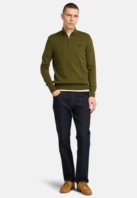Olivgrüner Reißverschluss-Pullover mit gerippten Bündchen und Saum, kombiniert mit dunklen Jeans und beigen Schuhen. Einfaches Design mit minimalen Branding-Details.