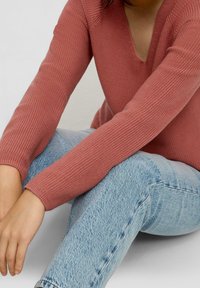 Rippiger, staubiger rosafarbener Strickpullover mit V-Ausschnitt; getragen mit hellblauen, eng anliegenden Jeans. Fokus auf Textur und Design des Stoffes und der Passform.