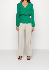 Blusa verde avvolgente con maniche lunghe, vita arricciata e scollatura a V profonda. Accompagnata da pantaloni beige larghi e tacchi neri con cinturini.