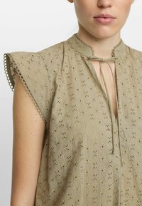 Ljus olivgrön blus med puffärmar, broderade detaljer, mandarin-krage och snörning fram. Strukturerad tyg med ögla-mönster.