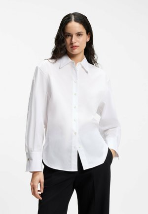 Femme aux longs cheveux foncés portant une chemise blanche boutonnée et un pantalon noir, debout avec une main dans la poche devant un fond uni.