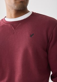 Man wearing a maroon crewneck sweatshirt over a white t-shirt, featuring a small black bird logo on the chest.