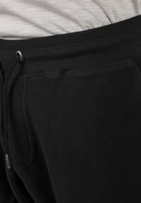 Pantalon de survêtement noir en tissu texturé, doté d'une taille élastique à cordon et d'une poche sur le côté, partiellement visible au-dessus d'un haut gris clair.