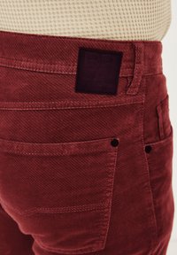 Rote Cordhosen mit strukturierter Oberfläche, ausgestattet mit einem dunklen Etikett auf dem Bund und sichtbaren Taschen mit kontrastierender Naht.