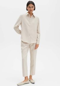 Camicia beige con bottoni e colletto, abbinata a pantaloni sartoriali coordinati. Scarpe basse e morbide con texture trapuntata completano l'outfit.