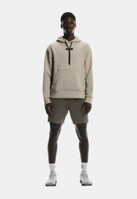 Haut à capuche beige avec une poche, cordons de serrage noirs, associé à un short marron. Le modèle porte des chaussures blanches avec des accents noirs et des chaussettes blanches.