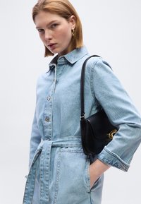 Νέα γυναίκα με κοντά ξανθά μαλλιά, που φοράει ανοιχτό τζιν φόρεμα με κουμπιά και ζώνη, κρατώντας μικρή μαύρη δερμάτινη τσάντα ώμου.