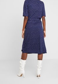 Kobieta w granatowej sukience midi z drobnym kwiatowym wzorem i białych, luźnych botkach na obcasie, stojąca na gładkim białym tle.