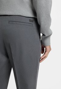 Gray trousers with a textured fabric, featuring a back pocket and a small logo accent near the waistband. Close-up view of the side.