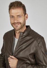 Braune Lederjacke mit einem Reißverschluss vorne, gesteppten Schulterakzenten und einem runden Logo-Patch auf dem linken Ärmel. Glatte Textur.