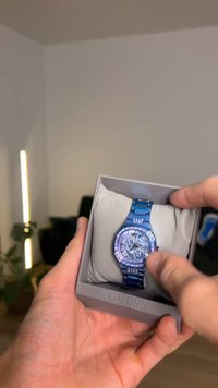 Modra zapestna ura s prozornim številčnico, okrašena z dragulji, kovinskim paščkom, predstavljena v sivi škatli s mehko notranjo blazino. Prikazuje kazalce in podrobnosti.