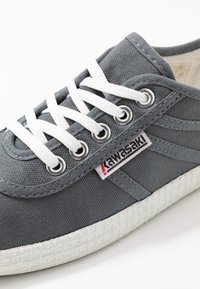 Sneaker grigio in tela con lacci bianchi, occhielli metallici e suola bianca testurizzata. Presenta un'etichetta con il marchio sul lato, con dettagli rossi.