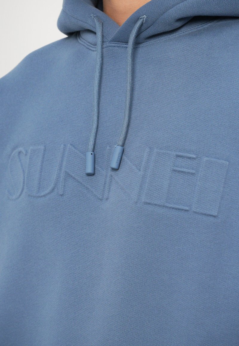 Felpa con cappuccio di colore azzurro chiaro, realizzata in tessuto morbido. Presenta un discreto logo "SUNNEI" in rilievo e punte del cordino abbinate.