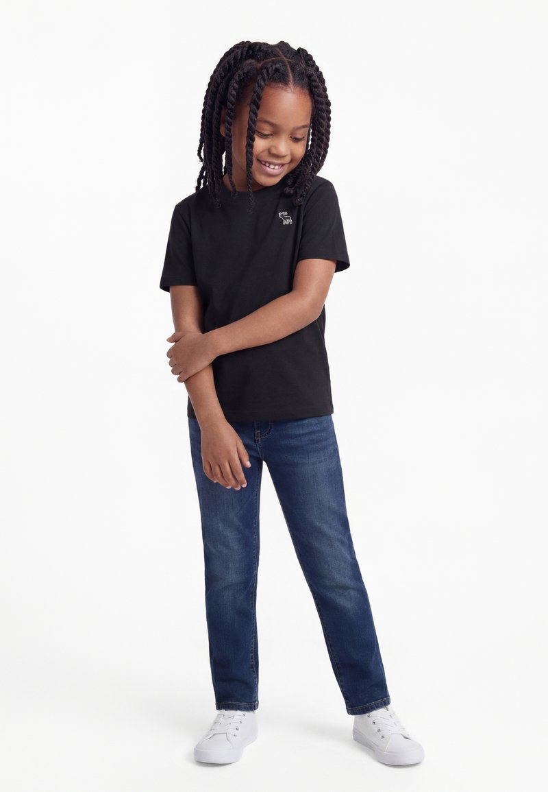 Enfant aux cheveux tressés portant un t-shirt noir, un jean bleu et des baskets blanches, debout les bras croisés, regardant vers le bas et souriant.