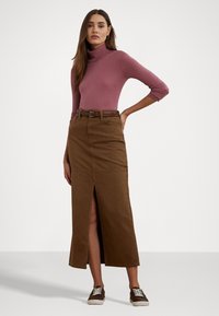 Hnedá denimová maxi sukňa s predným rozparkom; spárovaná s priliehavým, svetloružovým rolákom. Hnedé tenisky dotvárajú outfit.