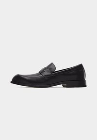 Mocassin pour homme en cuir noir avec talon bas, bout arrondi, et bride décorative sur le dessus, présenté de profil sur un fond uni.