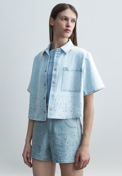 Jeune femme portant une veste en jean bleu clair à manches courtes et un short assorti ornés de sequins, debout devant un fond neutre.