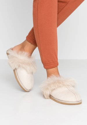Beige falskinnslipper med en myk, fluffy mansjett. Overdelen er laget av semsket skinn, med en avrundet tå og slitesterk gummisåle.