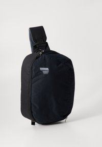 Black sling bag with a structured shape, sturdy fabric, adjustable strap, and a prominent logo detail in light gray. Textured accents on strap.