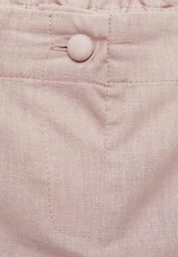Světle růžová lněná látka s detailem knoflíku a prošitou kapsou. Textura je jemná a mírně hrubá, což zdůrazňuje přírodní materiál.