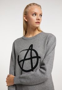 Szürke kerek nyakú pulóver, amely elülső részén egy nagy fekete anarchia szimbólum mintázattal készült. Puha kötött anyagból készült.