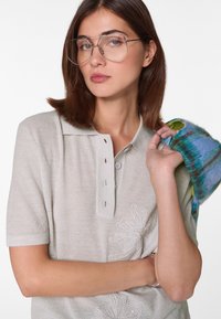 Lichtgrijze gebreide poloshirt met een gestructureerde afwerking, geborduurd bloemenmotief en knoopsluiting. Een veelkleurig gebreid accessoire hangt over één arm.