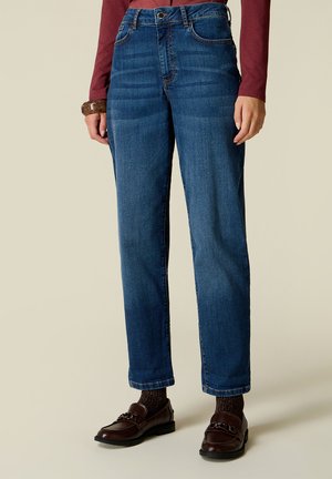 Frau trägt blaue Jeans mit geradem Bein, braune Loafers, dunkel gemusterte Socken, ein bordeauxfarbenes Langarmshirt und ein braunes Armband.