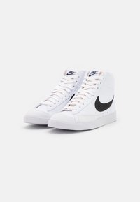 Białe sneakersy typu high-top z czarnym zamszowym logo Nike, sznurowane, z gumową podeszwą i teksturowanymi akcentami z tkaniny. Styl minimalistyczny.