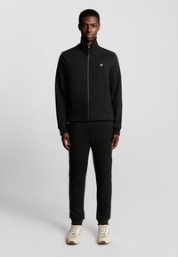 Zwarte zip-up sweatshirt en jogger set gemaakt van zachte stof. Bevat een ritssluiting, hoge kraag en een klein logo op de borst. Eenvoudig ontwerp.