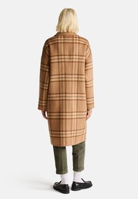 Brązowy płaszcz w kratę, wykonany z miękkiego materiału, o luźnym kroju, z długimi rękawami i kołnierzem. W zestawie z zielonymi spodniami kordrowymi i czarnymi butami.