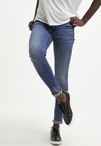 Modré džíny skinny s mírným vyblednutím a ohrnutými manžetami, doplněné černými šněrovacími teniskami na světlém pozadí.