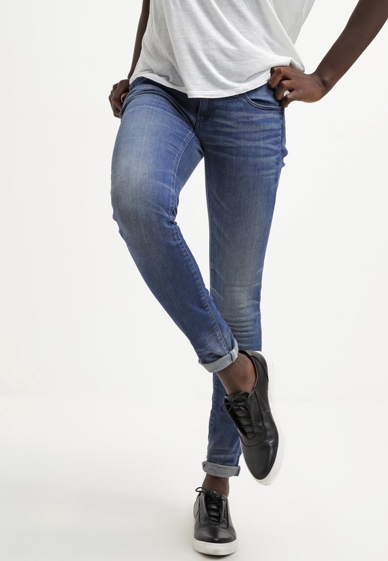 Modré džíny skinny s mírným vyblednutím a ohrnutými manžetami, doplněné černými šněrovacími teniskami na světlém pozadí.