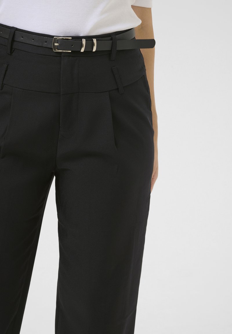 Sección media de una persona que lleva pantalones plisados negros con cinturón negro y una camiseta blanca de manga corta acanalada sobre un fondo liso.