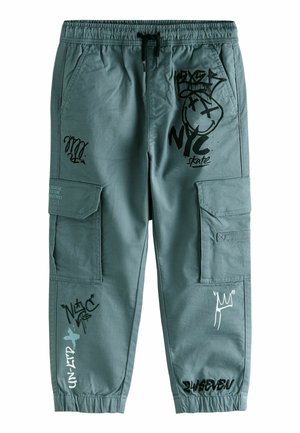 Pantalon cargo bleu sarcelle avec taille et poignets élastiques, avec des textes et motifs graphiques style graffiti en noir et blanc sur les jambes avant et les poches.