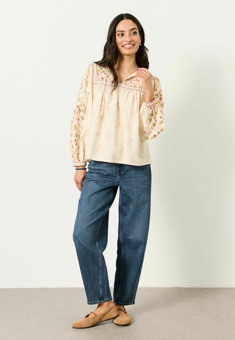 Mujer sonriendo, con una blusa crema bordada con flores y mangas abullonadas, jeans anchos azules y zapatos planos color canela, de pie frente a una pared blanca.