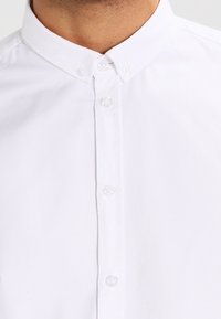 Camicia bianca con colletto, caratterizzata da una trama liscia e bottoni in plastica trasparente. La vista ravvicinata mostra il tessuto e i dettagli della cucitura.