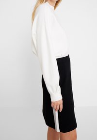 Maglione bianco a maniche lunghe con una vestibilità ampia, abbinato a una gonna a matita nera con una striscia verticale bianca lungo il lato.