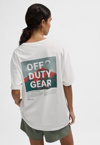 Vit oversized t-shirt med ett grafiskt tryck på baksidan, med texten "OFF DUTY GEAR", i gröna och röda färger samt en texturerad bergillustration.