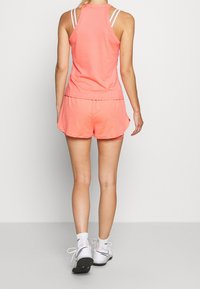 Růžový sportovní top bez rukávů s designem racerek, spárovaný s odpovídajícími texturovanými šortkami. Model nosí bílé tenisky s černými akcenty a bílé ponožky.