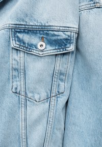 Τσέπη από ανοιχτό μπλε τζιν μπουφάν με καπάκι ασφαλισμένο με μεταλλικό κουμπί χαραγμένο με τη φράση "HUGO JEANS" και εμφανή πορτοκαλί ραφή.