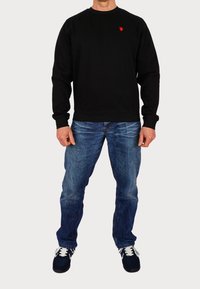 Svart sweatshirt med rund hals och röd logotyp, tillsammans med blå jeans. Modellen bär marinblå sneakers med vita detaljer.